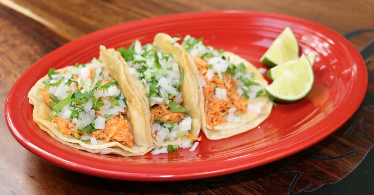 Three shredded chicken tacos topped with chopped onions and cilantro, served on soft corn tortillas with lime wedges on the side.