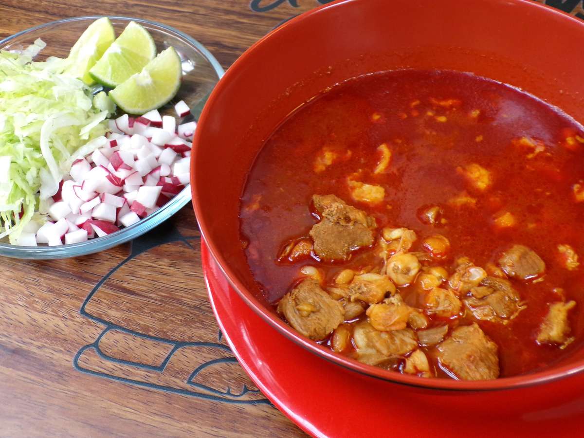 Bowl of red pozole served with lime, chopped onion, cilantro, jalapeños, and a shrimp cocktail in a michelada glass.
