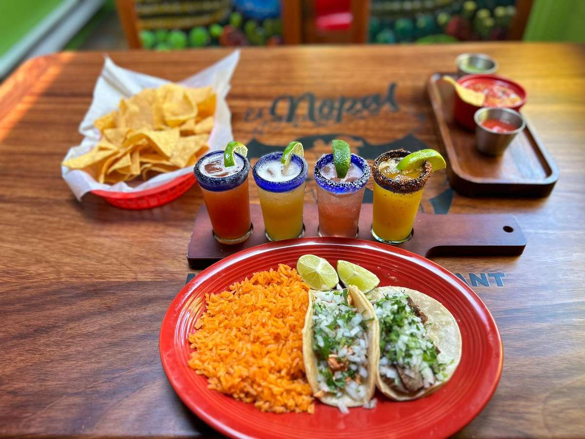 Tacos with rice, lime wedges, chips with salsa, and a flight of four colorful margaritas garnished with salt rims and limes.
