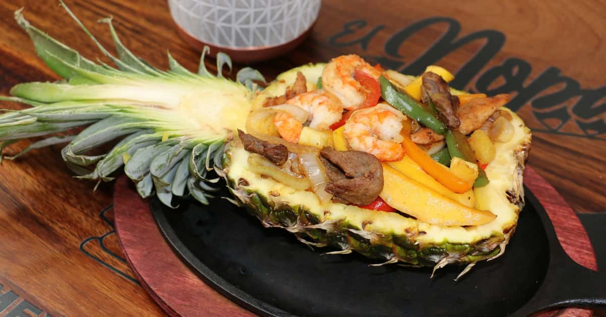 Grilled shrimp, beef, chicken, and bell peppers served in a halved pineapple bowl on a sizzling platter.