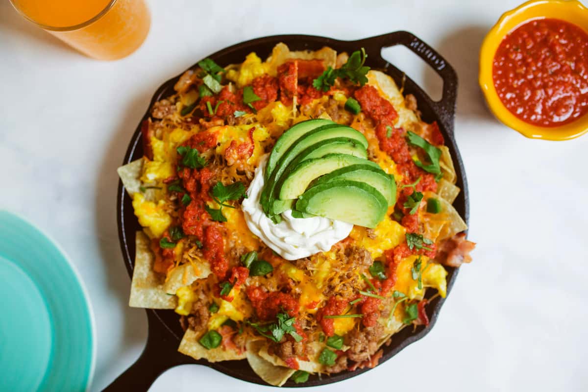 Loaded breakfast nachos topped with scrambled eggs, ground sausage, cheddar cheese, red salsa, sour cream, and avocado slices, served in a cast iron skillet.