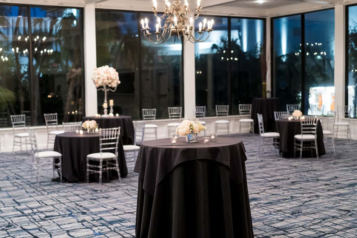 Modern waterfront cocktail reception room with black linen tables, floral centerpieces, and floor-to-ceiling evening views