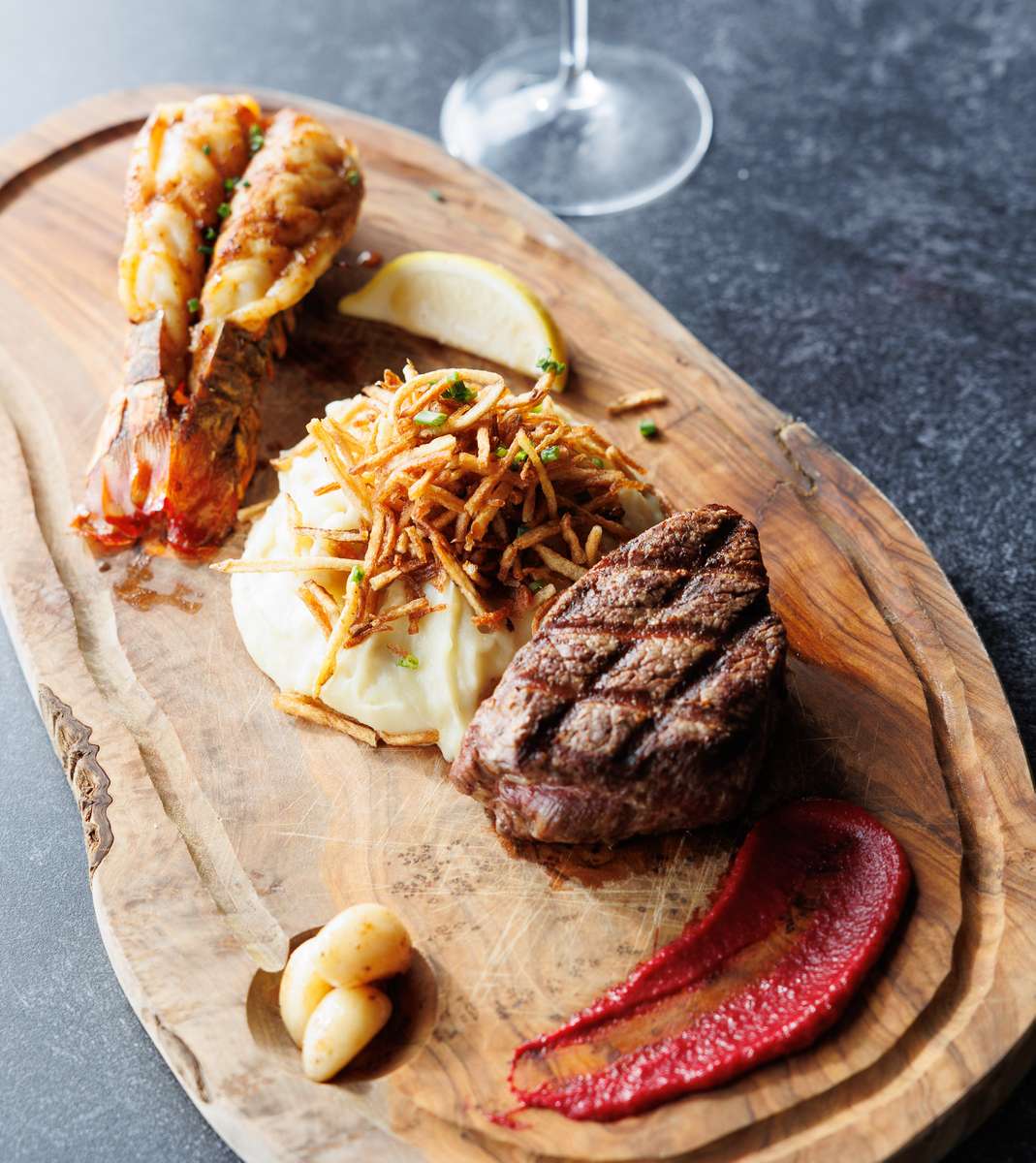 Grilled filet mignon and lobster tail served with creamy mashed potatoes and roasted garlic on a rustic wooden board
