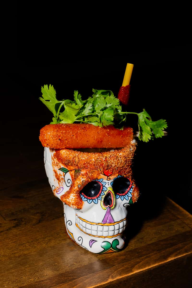 Spicy michelada-style cocktail served in a decorative skull mug with chili seasoning and fresh cilantro garnish