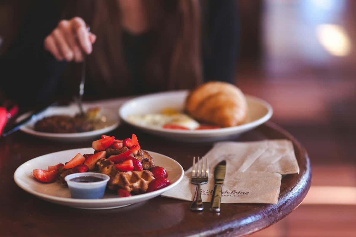 Breakfast scene with waffles topped with strawberries and syrup, a croissant with eggs, and a person eating in a cozy café setting.