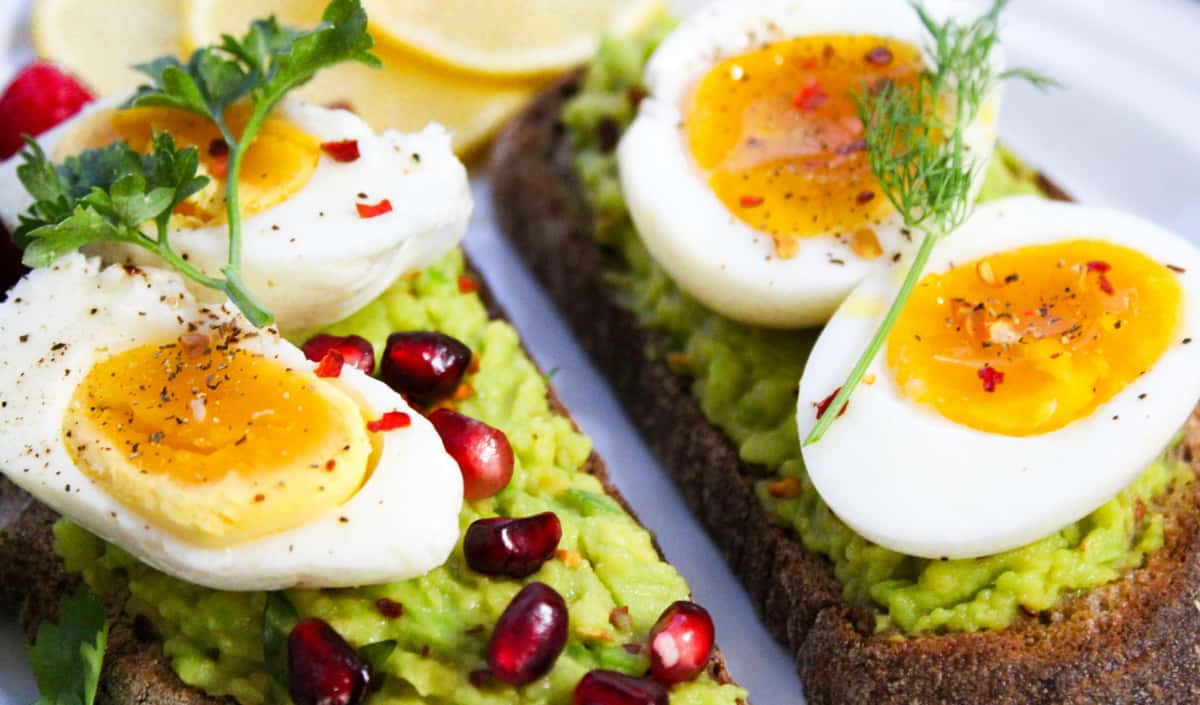 Gourmet avocado toast topped with perfectly soft-boiled eggs, pomegranate seeds, and fresh herbs on whole grain bread, garnished with cracked black pepper and chili flakes for a vibrant and nutritious breakfast.