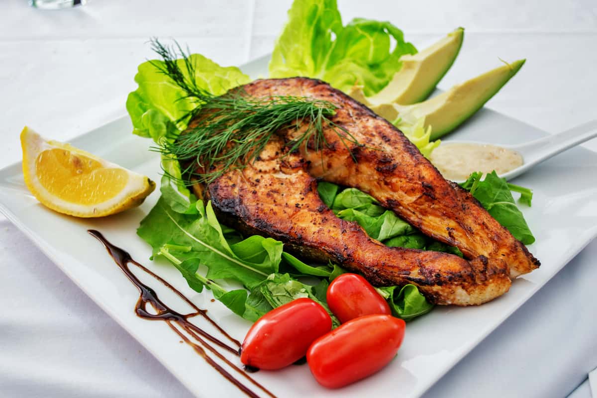 Grilled salmon steak served on a bed of fresh greens with cherry tomatoes, avocado slices, lemon wedge, and balsamic drizzle.