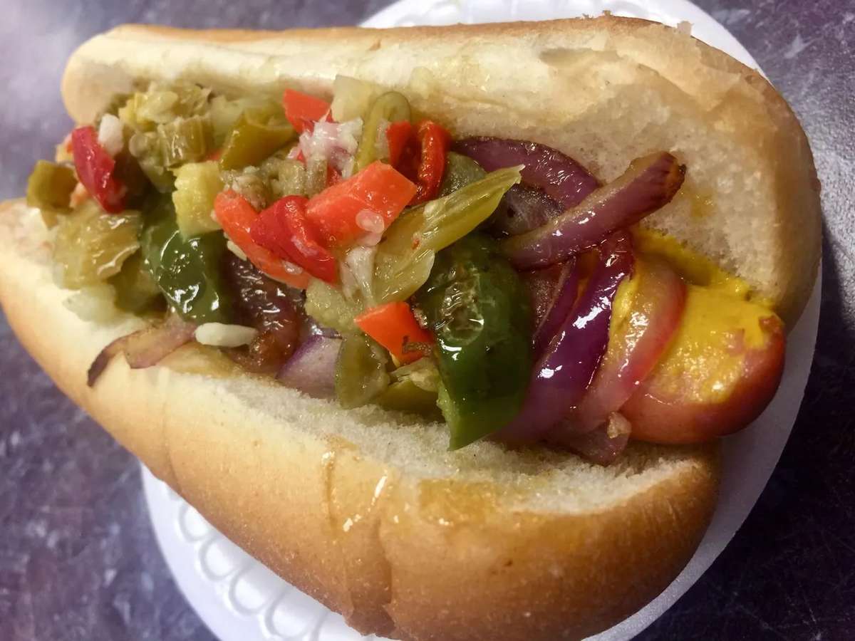 Grilled hot dog topped with sautéed onions, mustard, and a colorful mix of giardiniera peppers in a fresh bun.