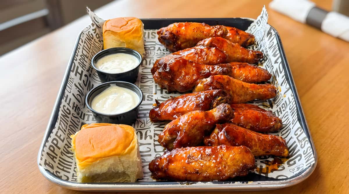 Tray of glossy red-orange chicken wings with two small cups of white dipping sauce and two dinner rolls on patterned paper.