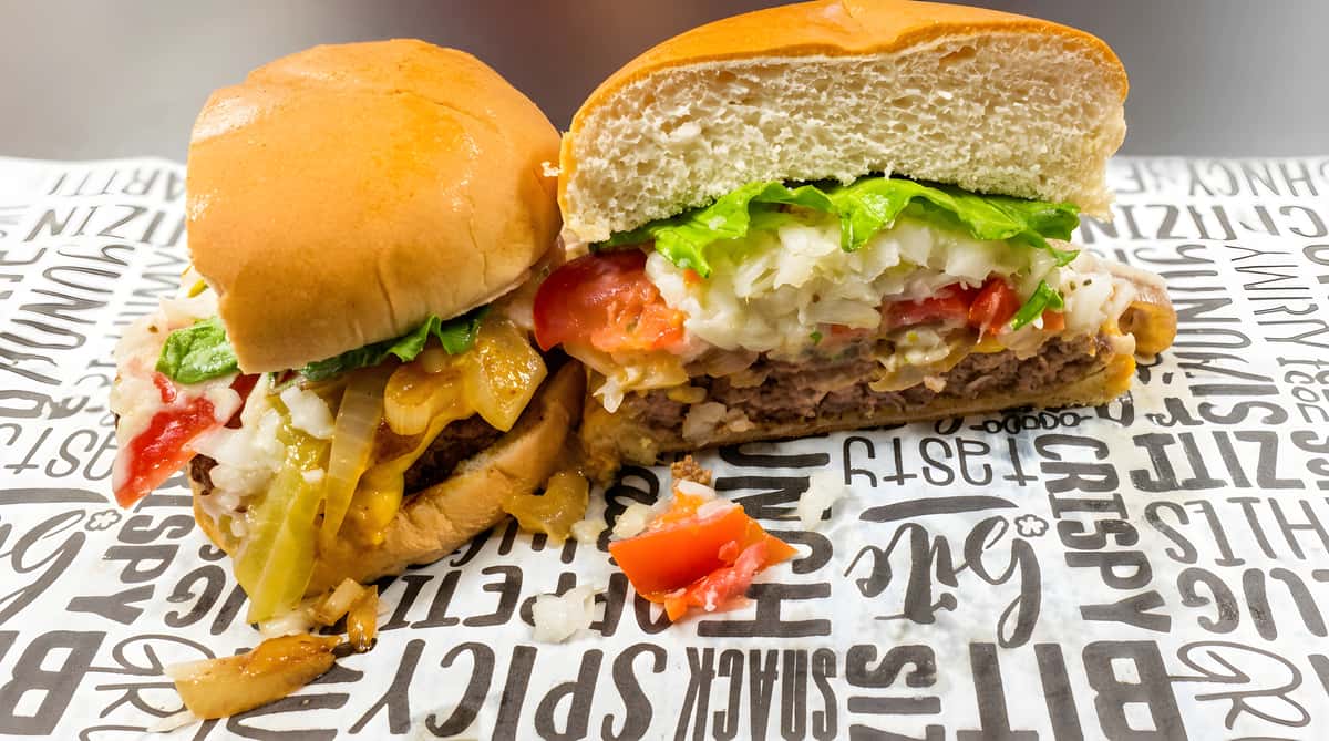 Burger cut in half on a wrapper, showing a beef patty, melted cheese, sautéed onions, lettuce, and tomatoes.