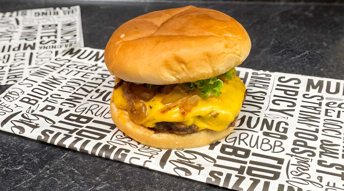 Cheeseburger with melted yellow cheese, caramelized onions, and lettuce on a bun, resting on patterned paper.