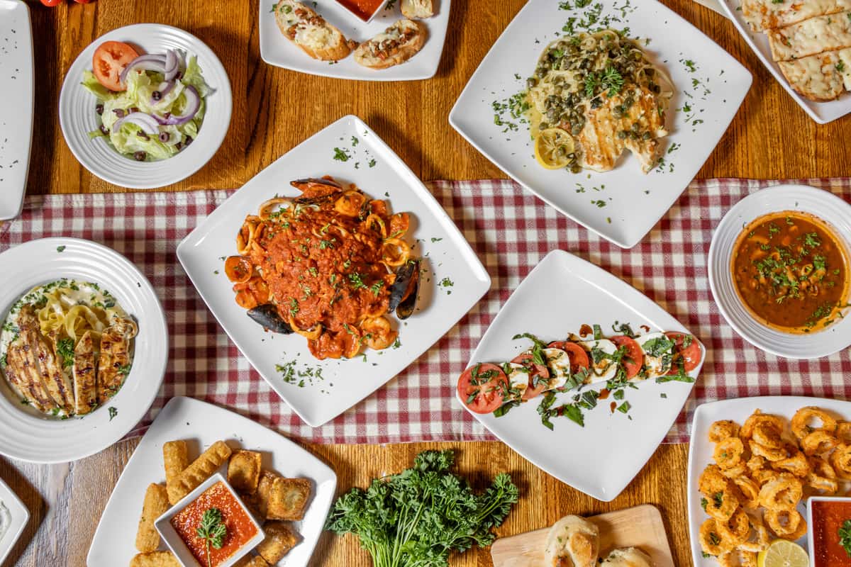 Overhead view of Italian restaurant spread with seafood pasta in tomato sauce, caprese salad with fresh mozzarella and basil, creamy pasta with grilled chicken, garlic bread, seasoned fried calamari, mixed green salad, soup, and bruschetta on a checkered tablecloth.