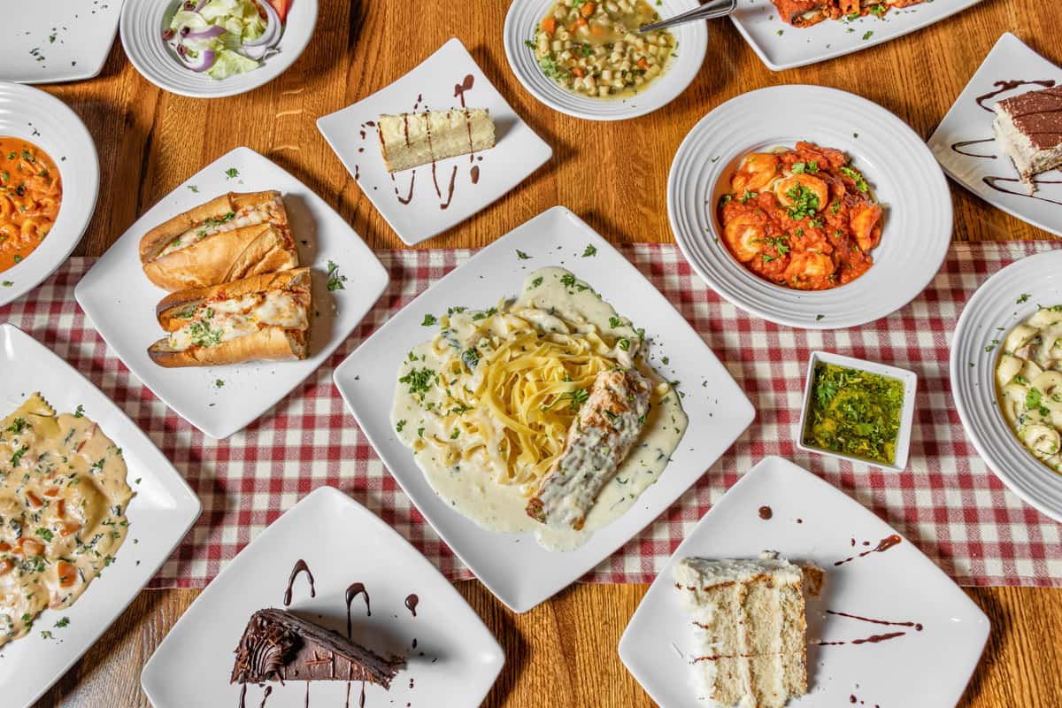 Top-down view of an Italian restaurant table featuring creamy fettuccine Alfredo with grilled chicken, baked sub sandwiches, cheese ravioli in tomato sauce, assorted cakes and tiramisu, hearty vegetable soup, and fresh garden salad on a checkered tablecloth.