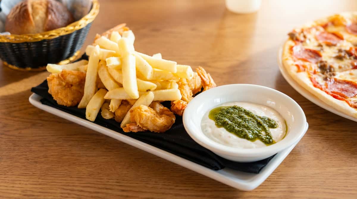 Fried shrimp and french fries on a black plate with a side of white sauce drizzled with green pesto.