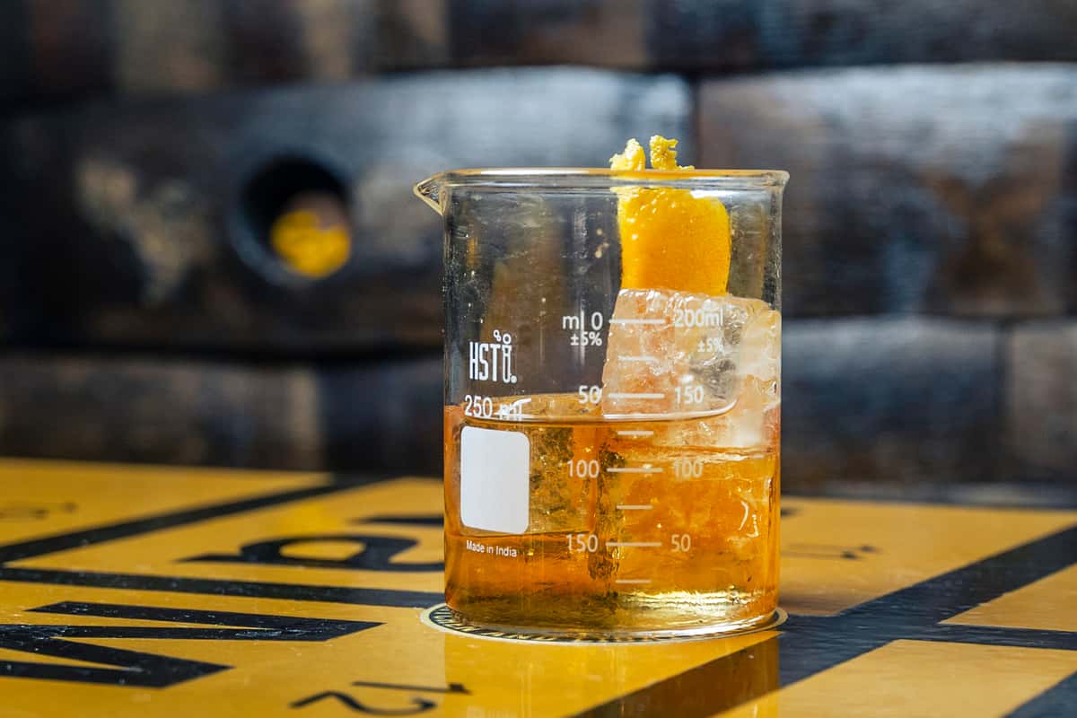 Photo of the Olde School House cocktail, served in a scientific beaker and garnished with an orange peel. Photo Credit: The Bacyard.
