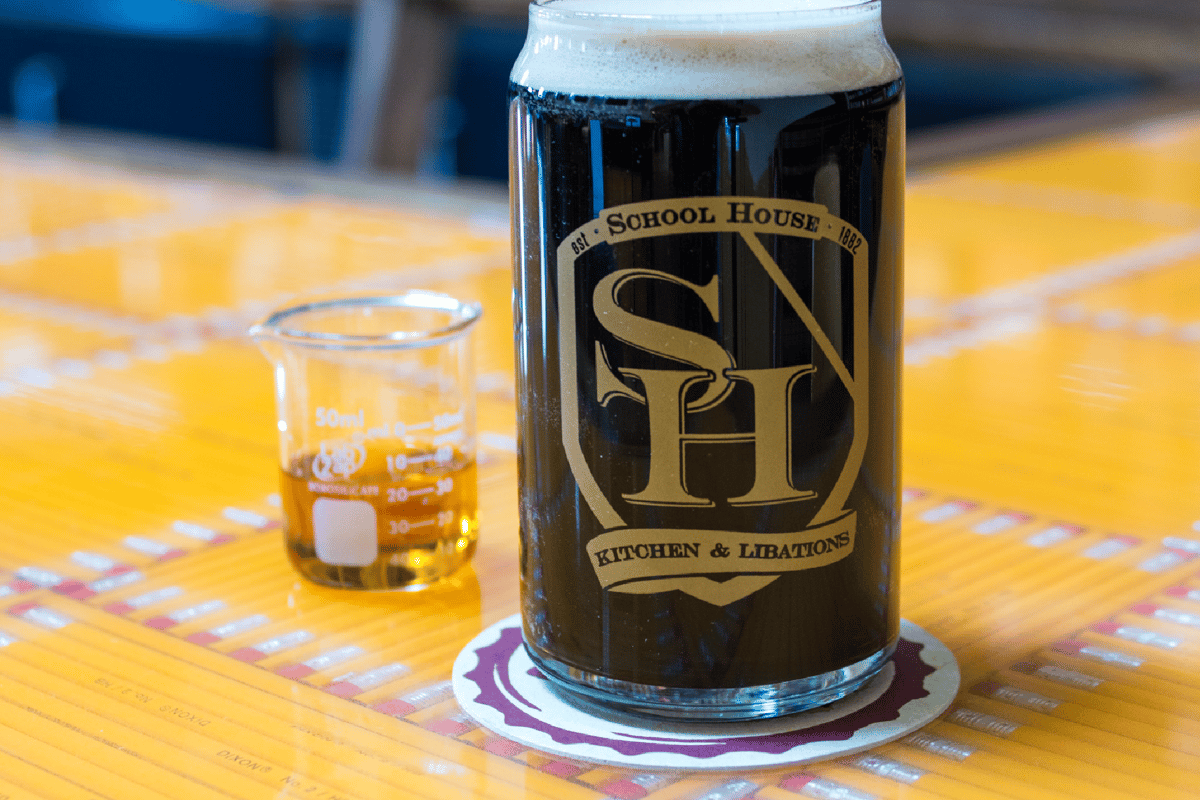 Photo of a dark beer in glass with a School House logo, with a shot of whiskey in a beaker
