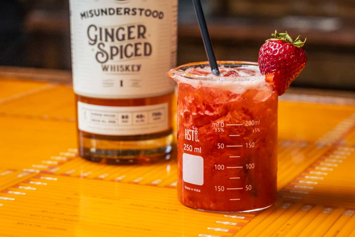 Photo of the Ms. Understood cocktail, served in a scientific beaker and garnished with a fresh strawberry.  A bottle of Misunderstood Ginger Spiced Whiskey sits in the background. Photo Credit: The Bacyard. 