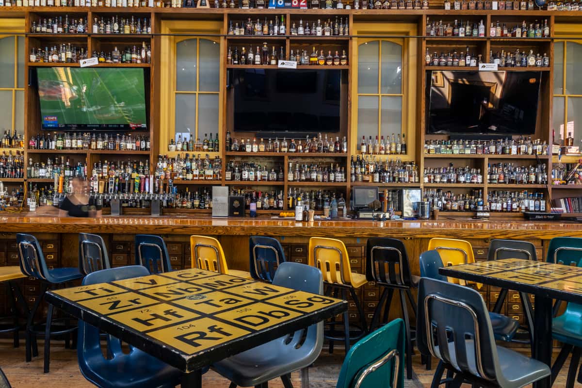 Photo of the "Library Bar" seating area with high-top tables decorated to look like the Periodic Table of Elements, school chairs converted into bar stools, and 2-story high shelving, filled with whiskey bottles, in the background. Photo Credit: The Bacyard.