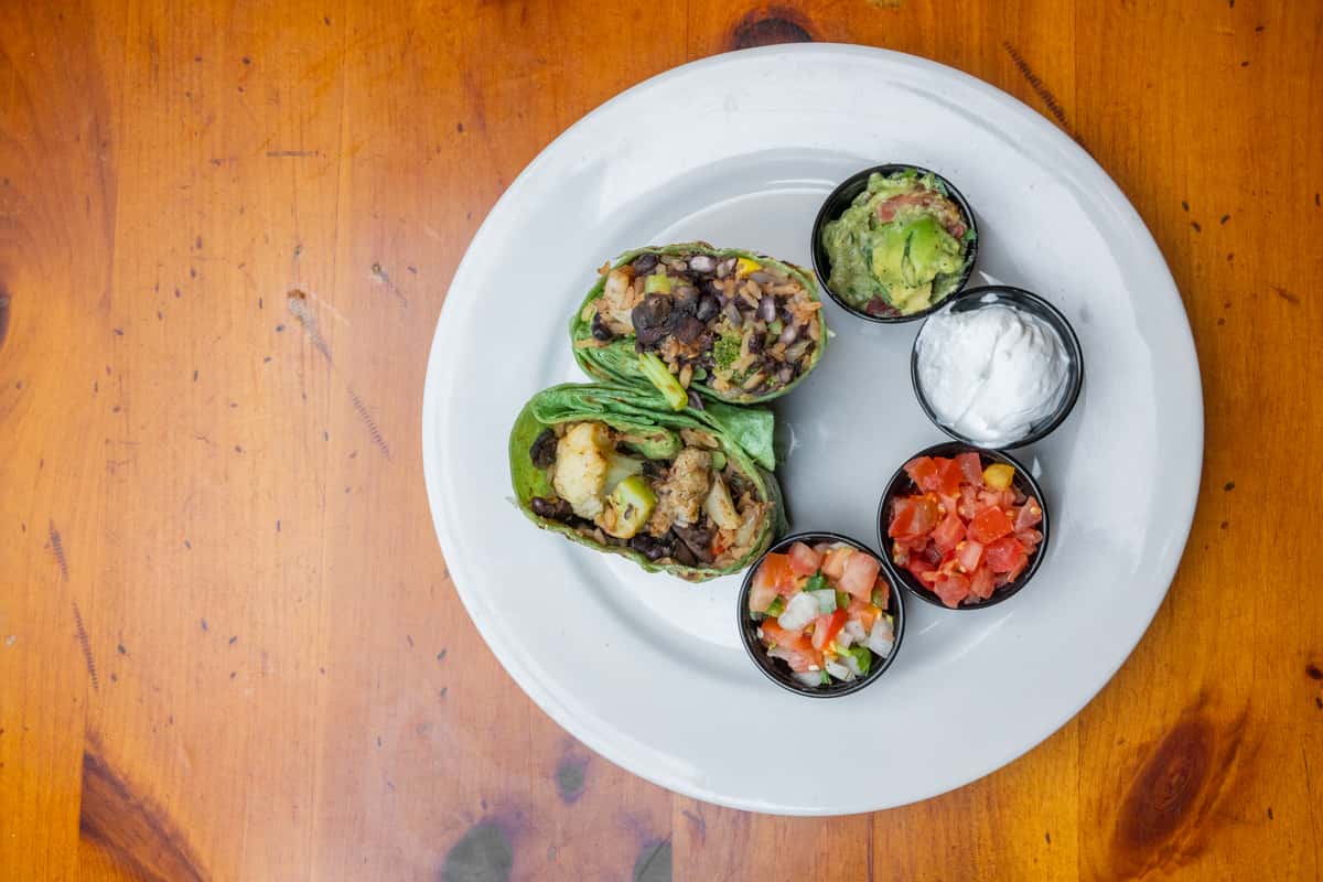 Overhead view of a fresh spinach wrap cut in half, filled with grilled chicken, black beans, rice, and vegetables, served with sides of guacamole, sour cream, pico de gallo, and diced tomatoes on a white plate over a rustic wooden table.