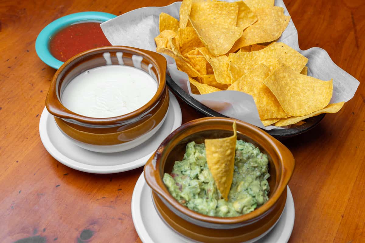 Crispy tortilla chips served with creamy white queso dip, fresh house-made guacamole, and classic red salsa in rustic bowls, showcasing a shareable Tex-Mex appetizer spread on a warm wooden table at a vibrant Mexican cantina.