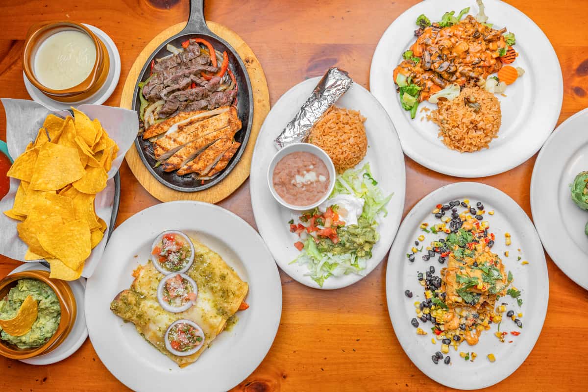 Mexican restaurant table spread featuring sizzling chicken and steak fajitas in a cast-iron skillet, cheesy enchiladas topped with salsa and onions, a burrito with Mexican rice and refried beans, loaded seafood salad with rice, and crispy tortilla chips with fresh guacamole and queso dip on a rustic wooden table, showcasing authentic Tex-Mex cuisine and vibrant dining ambiance.