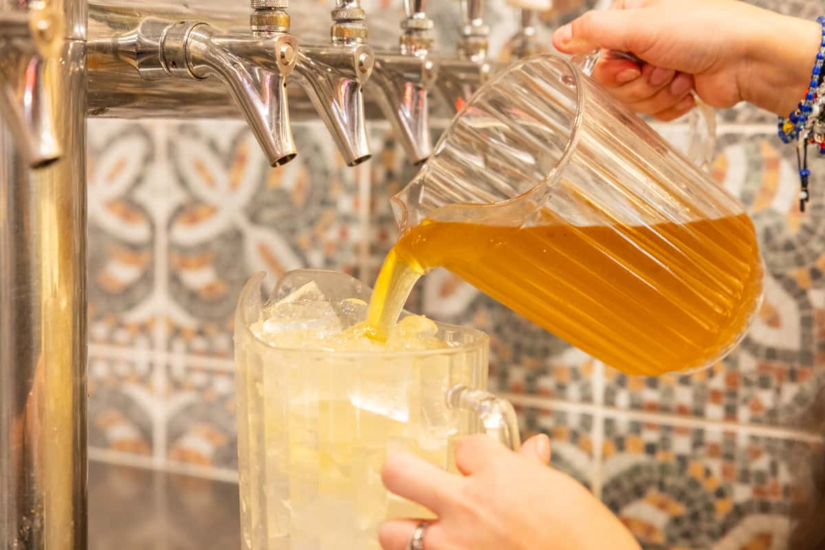 Draft beer being poured from stainless steel taps into a frosty glass mug filled with ice, capturing a refreshing golden beverage in a vibrant Mexican cantina bar setting with decorative tile backdrop and casual restaurant ambiance.