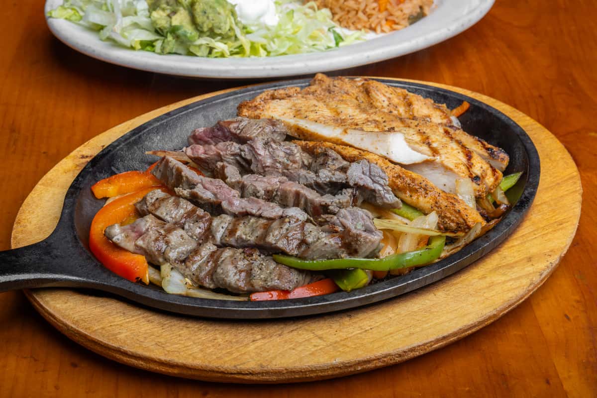 Sizzling chicken and steak fajitas served on a cast-iron skillet with grilled onions and bell peppers, presented on a wooden trivet with Mexican rice, guacamole salad, and sour cream in the background, highlighting authentic Tex-Mex cuisine in a vibrant cantina setting.