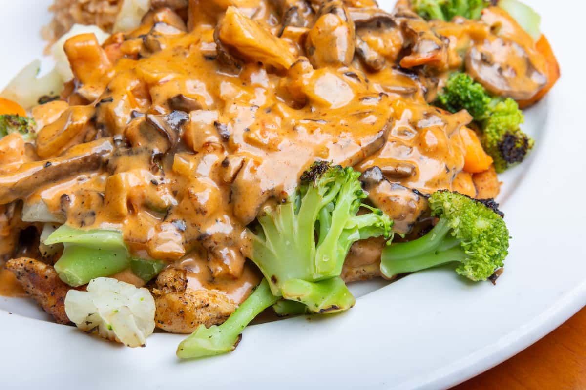 Grilled chicken breast topped with creamy mushroom sauce, served with steamed broccoli, carrots, and cauliflower on a white plate, showcasing a hearty American-style dinner with savory garlic cream sauce and fresh sautéed vegetables in a casual restaurant setting.