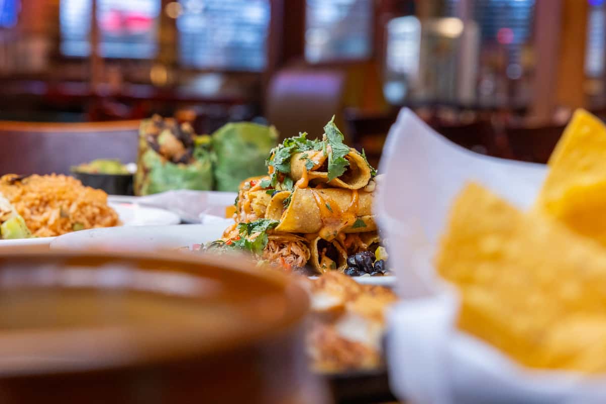 Crispy rolled chicken taquitos topped with fresh cilantro and sauce, served with Mexican rice, black beans, guacamole, and tortilla chips in a warm, casual Mexican restaurant setting.