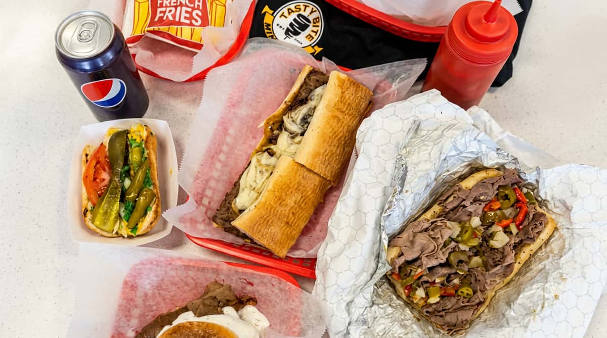 An array of fast food items: cheesesteak, hot dog with relish, Italian beef sandwich, fries, and a Pepsi cup.