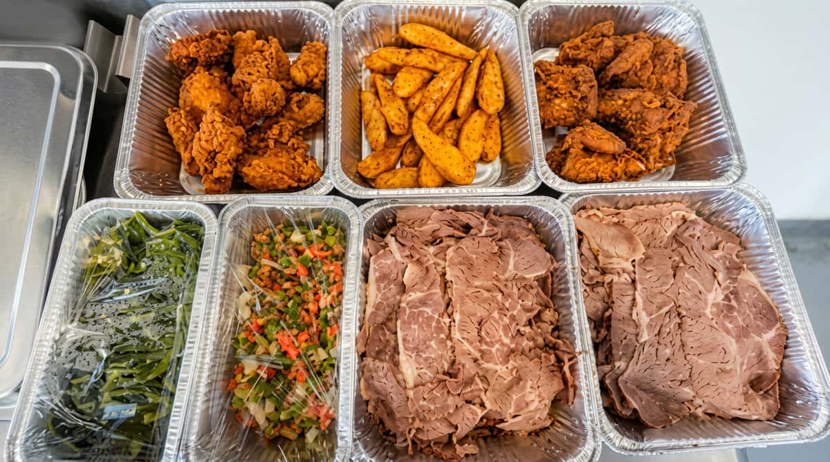 Six foil trays of food: fried chicken, potato wedges, seasoned dark meat, green vegetables, coleslaw, and sliced roast beef.