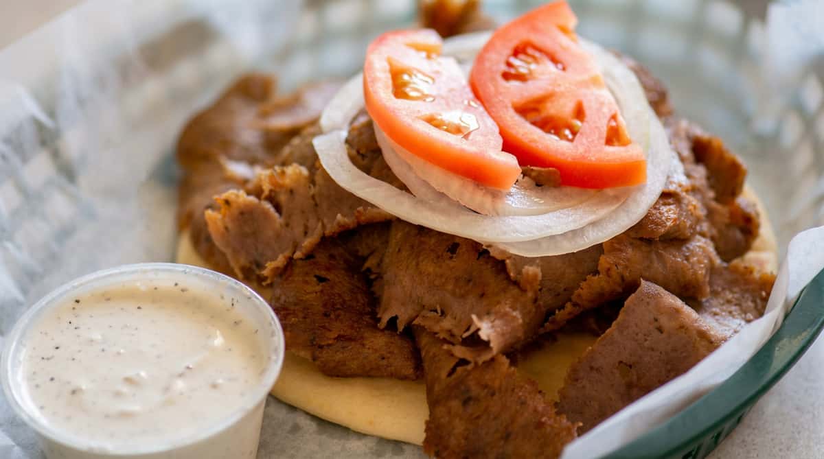 Gyro meat on pita bread topped with sliced tomatoes, raw onions, and a side of white sauce.