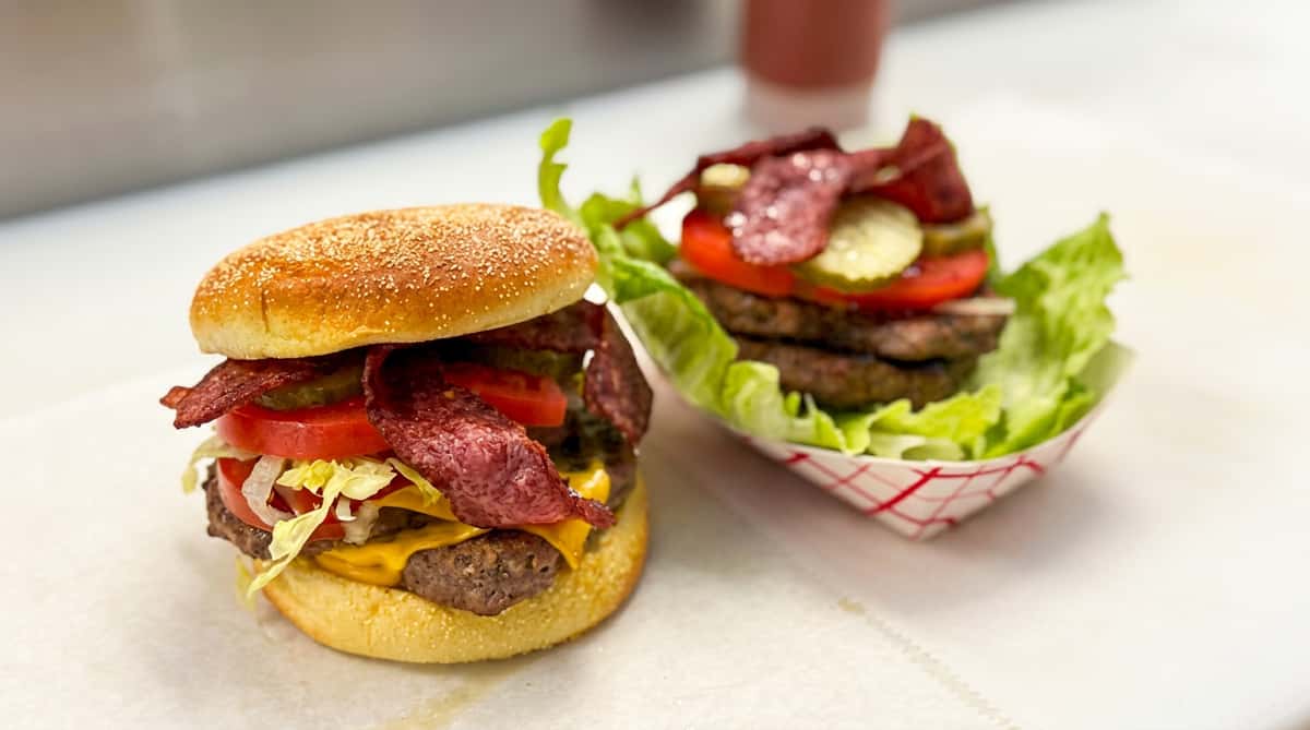 A bunned burger with cheese, bacon, and vegetables, next to a lettuce-wrapped burger with similar toppings.