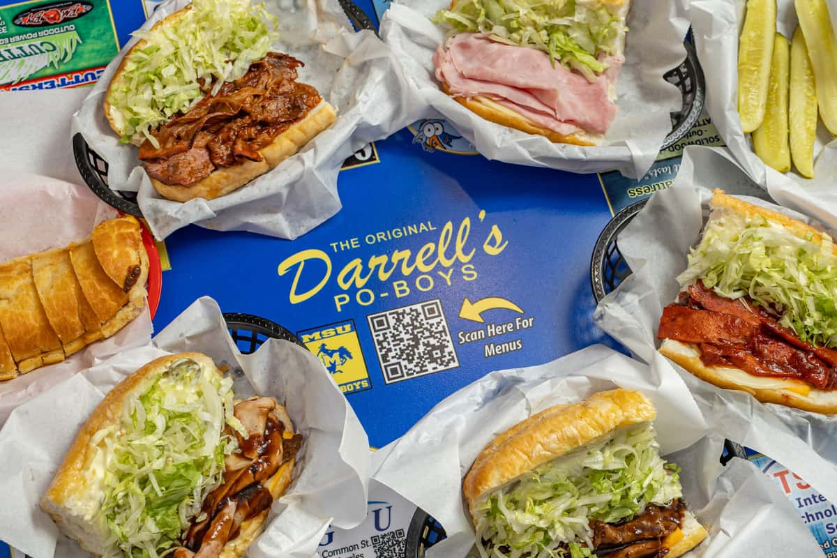 Close‑up of several po'boy sandwiches served in paper‑lined baskets on a blue tray branded with 'Darrell's Po‑Boys'. The sandwiches are in soft hoagie rolls, filled with shredded lettuce and various meats (roast beef gravy, sliced ham, pulled pork, bacon) and accompanied by crisp pickle spears at the edge.