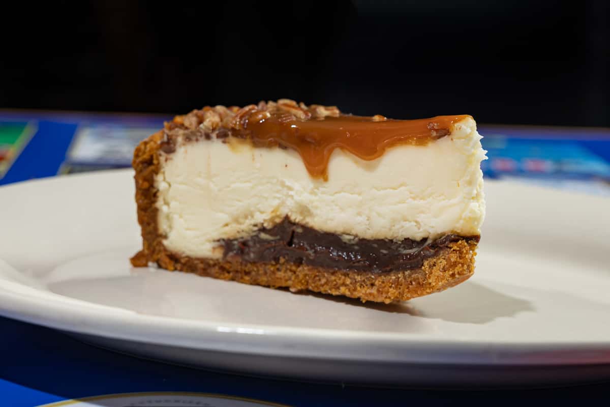 Slice of caramel pecan cheesecake served on a white plate, featuring a rich graham cracker crust, a layer of chocolate fudge, creamy cheesecake filling, and a thick caramel topping with chopped pecans.