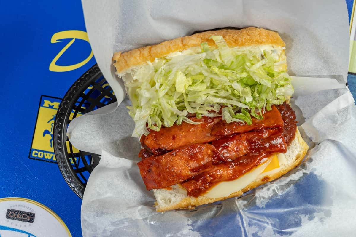 Open po'boy sandwich from Darrell's Po-Boys featuring grilled sausage links glazed in a tangy barbecue sauce, topped with shredded lettuce and melted cheese on a toasted hoagie roll, served in a paper-lined basket.