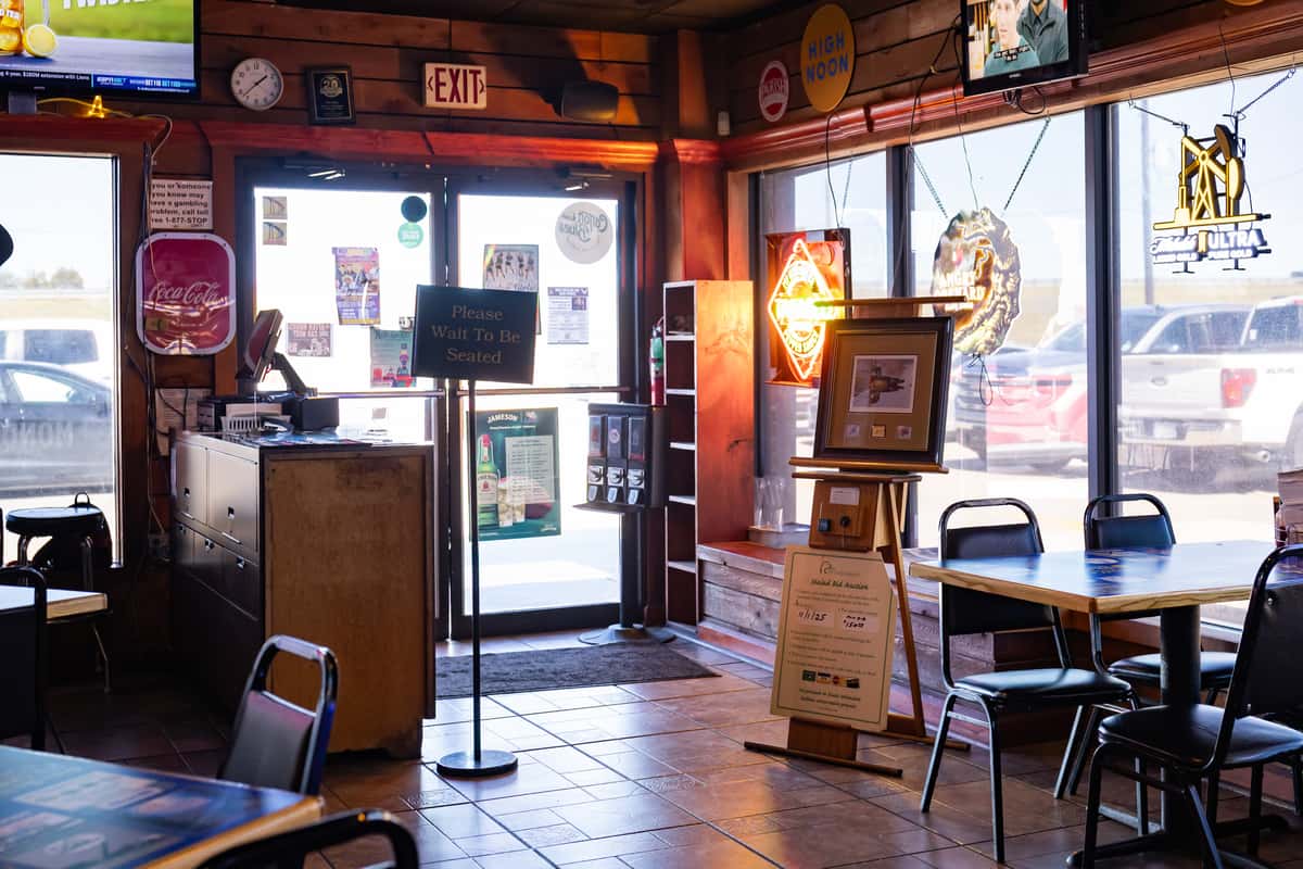 View of Darrell's restaurant entrance from inside, featuring a host stand, a 'Please Wait to Be Seated' sign, and window seating surrounded by neon beer signs and natural light from large front windows