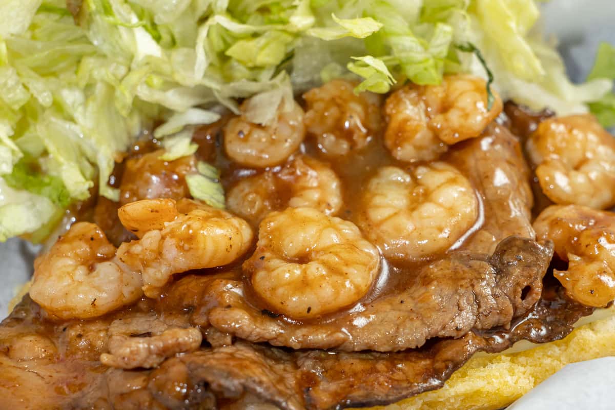Close-up of a surf and turf po'boy sandwich from Darrell's Po-Boys, featuring grilled steak and juicy Gulf shrimp smothered in rich brown gravy, topped with shredded lettuce and served on a toasted Louisiana-style roll.