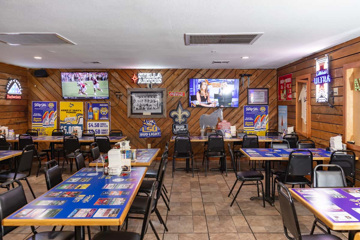 Interior of Darrell's restaurant showcasing a casual, sports bar-style dining area with blue tables, black chairs, and wood-paneled walls decorated with neon beer signs, LSU and Saints memorabilia, and flat-screen TVs playing sports and news. The space is clean, open, and family-friendly.