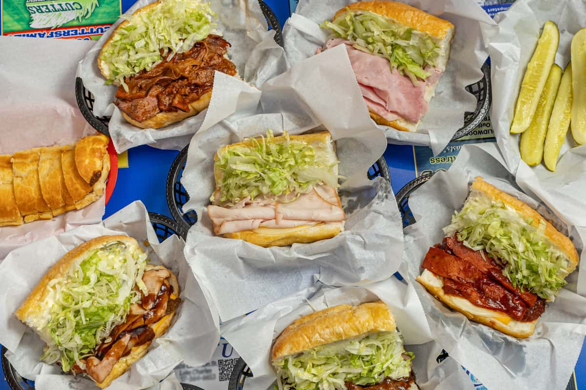 Selection of assorted po' boy sandwiches placed in baskets lined with parchment paper, featuring soft hoagie rolls filled with shredded lettuce and various meats — including pulled pork, sliced ham, smoked sausage, and bacon — arranged around a blue tray labeled 'The Original Darrell's Po‑Boys', accompanied by a side of pickle spears.