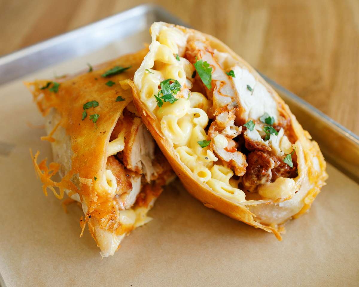 A golden-brown wrap, cut open to reveal crispy fried chicken and creamy macaroni and cheese, garnished with melted cheese and fresh parsley on parchment paper.