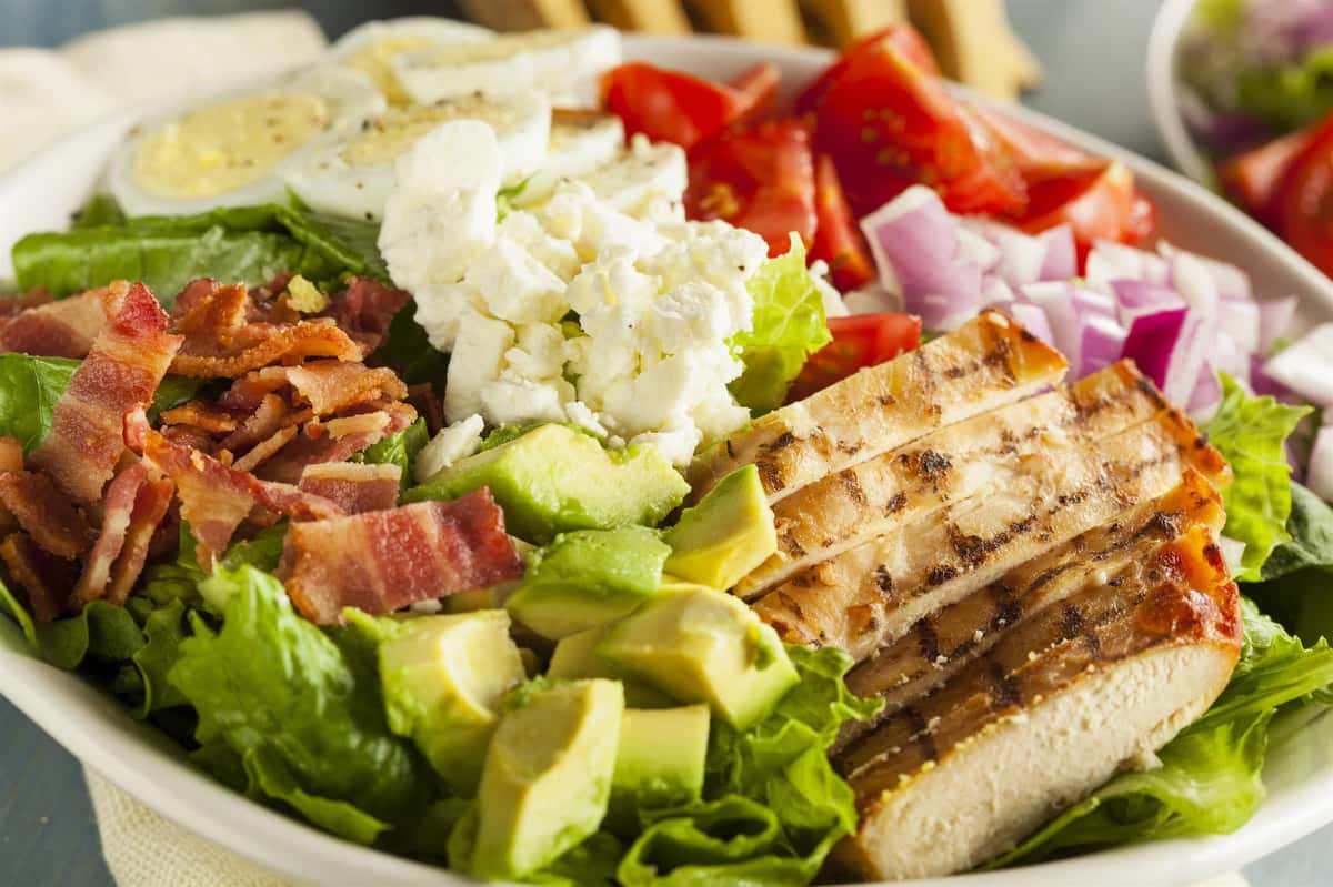 a bowl of salad with avacado, bacon, lettuce, chicken, tomato, onions, boiled eggs and cheese