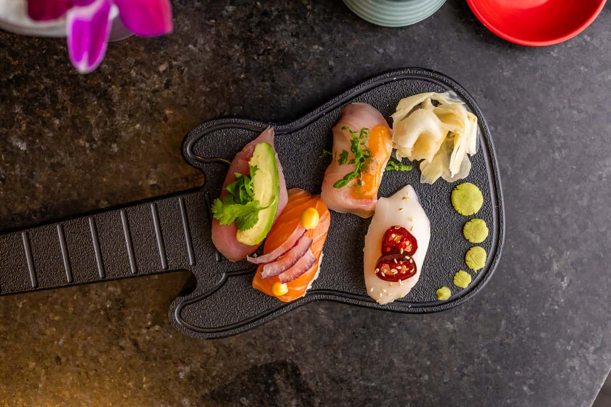 Assorted nigiri sushi with avocado, red onion, jalapeño, and herbs, served with wasabi and pickled ginger on a guitar-shaped plate.