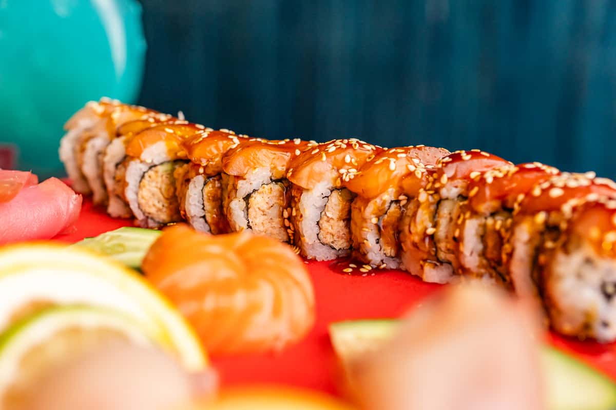 Close-up of a sushi roll topped with fresh salmon, drizzled with sauce, and sprinkled with sesame seeds on a vibrant red board.