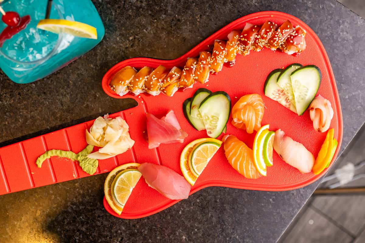 Assorted nigiri and sushi rolls with cucumber, lemon slices, wasabi, and pickled ginger on a red guitar-shaped serving board.