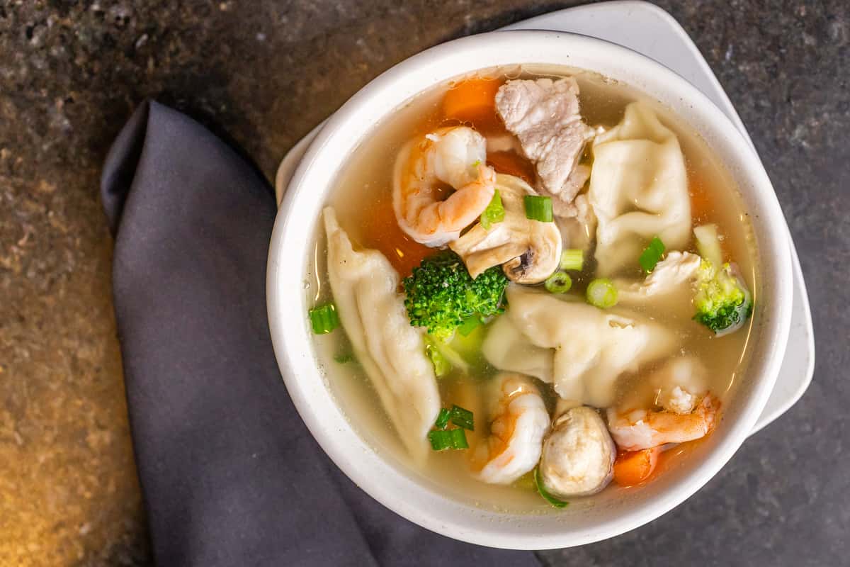 Clear broth dumpling soup with shrimp, pork slices, mushrooms, broccoli, carrots, and green onions in a white bowl.