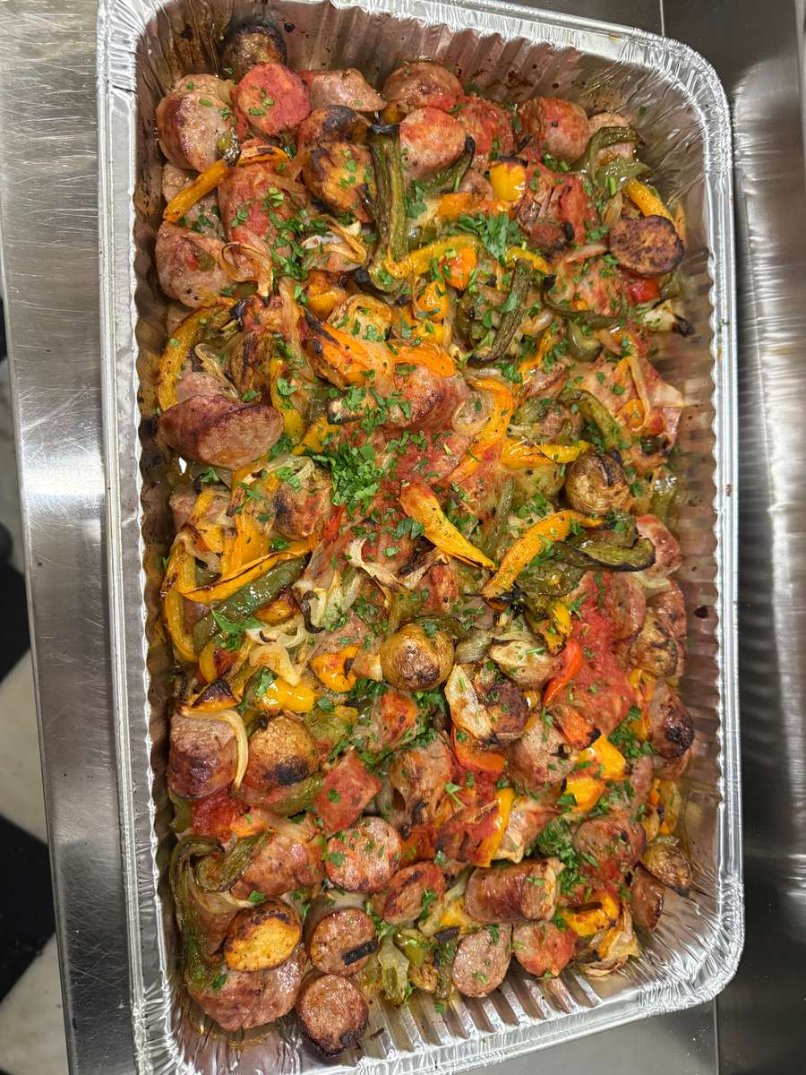 Tray of Italian sausage and peppers featuring grilled sausage slices, sautéed bell peppers, onions, and a light tomato sauce, garnished with fresh herbs—served in a foil pan for catering or family-style meals.