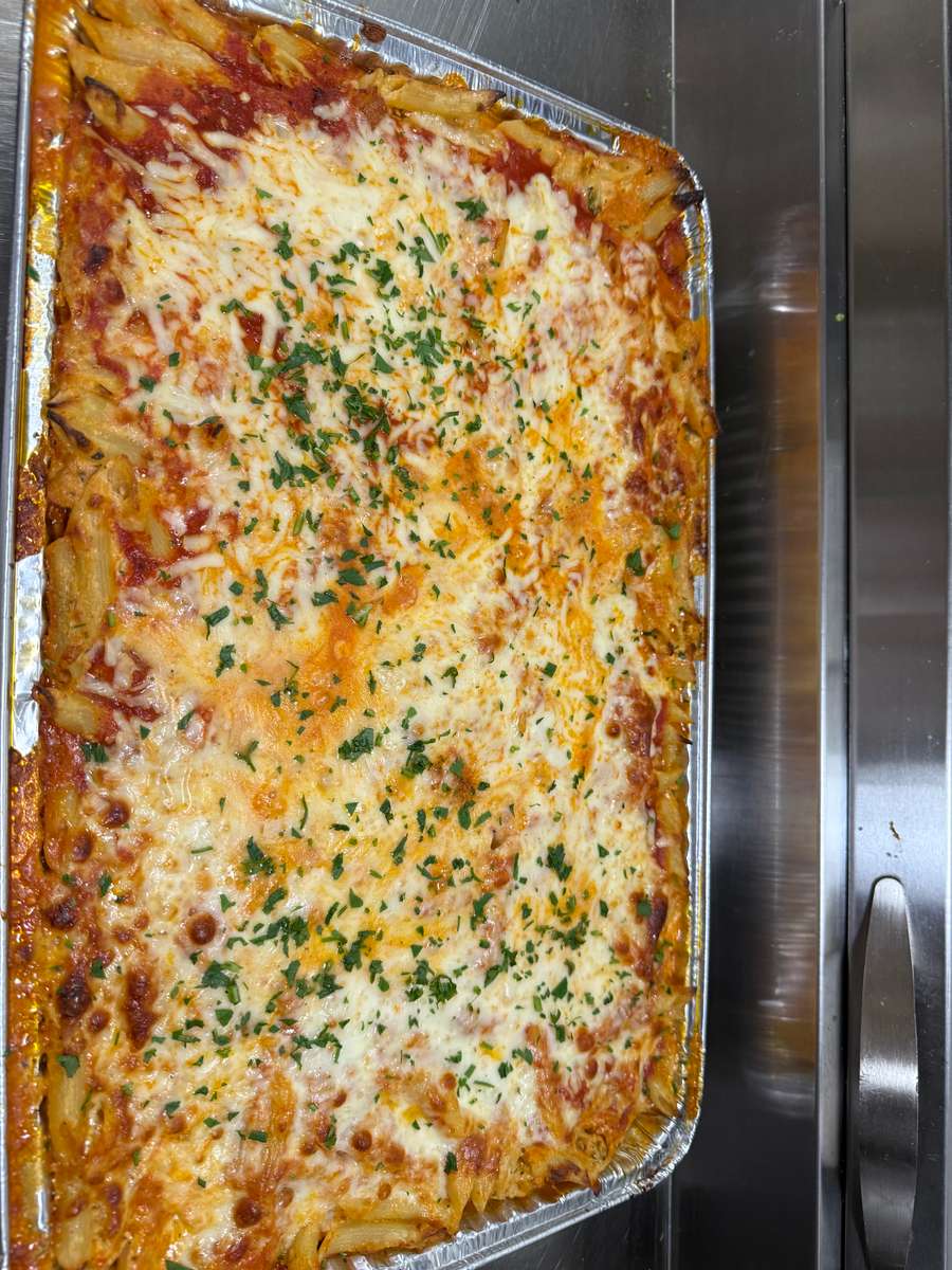 Overhead view of cheesy baked ziti pasta in a foil tray, layered with marinara sauce, melted mozzarella, and garnished with fresh chopped herbs—ideal for Italian catering or buffet service.