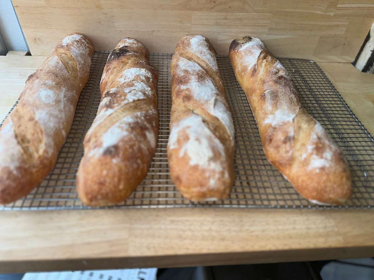 Four freshly baked artisan baguettes with golden crusts and rustic flour dusting, cooling on a wire rack atop a wooden counter in a bakery setting.