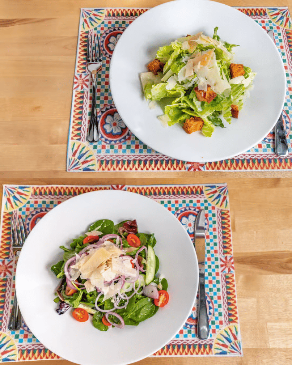 Overhead view of two fresh salads served on colorful patterned placemats—one classic Caesar salad with romaine lettuce, croutons, and shaved Parmesan, and one garden salad with mixed greens, cherry tomatoes, cucumber, red onion, and shaved cheese, plated on a light wooden table.